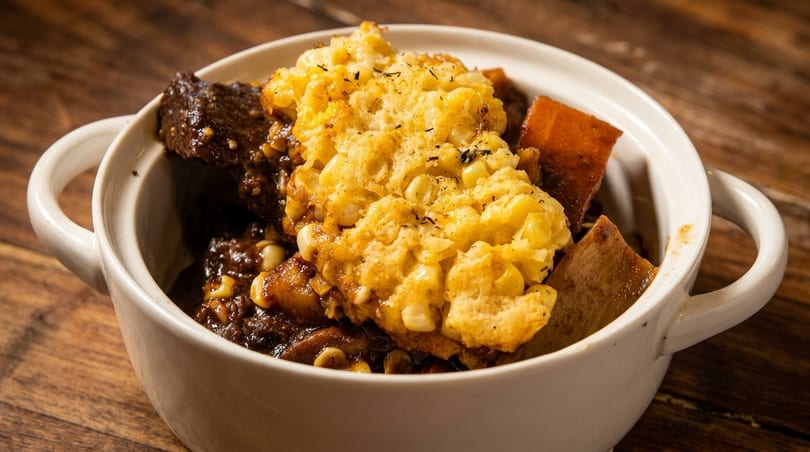 Hearty stew with dark meat, sweet potatoes, and a corn cobbler topping in a white bowl.