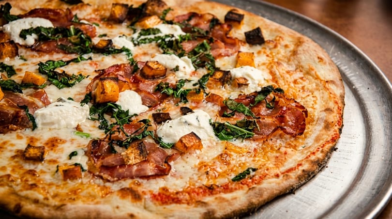 Close-up of a pizza with bacon, butternut squash, ricotta cheese, and greens on a metal tray.