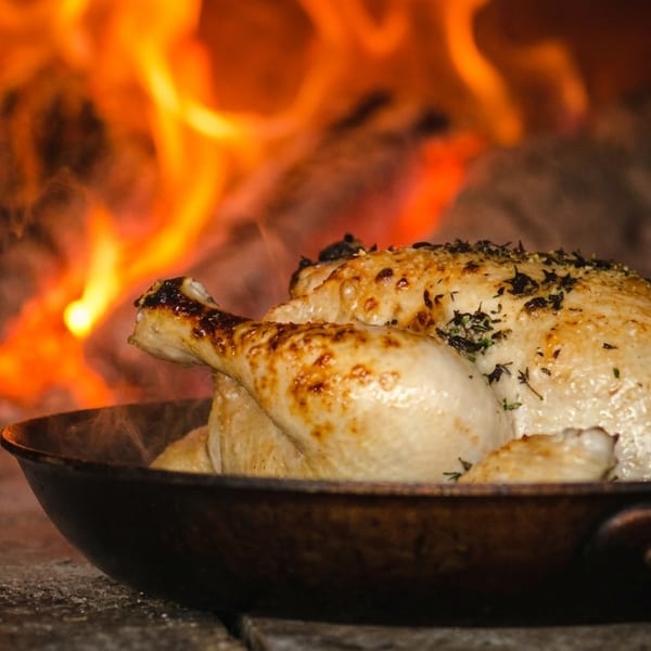Whole roasted chicken in a pan, cooking in a wood-fired oven with visible flames.