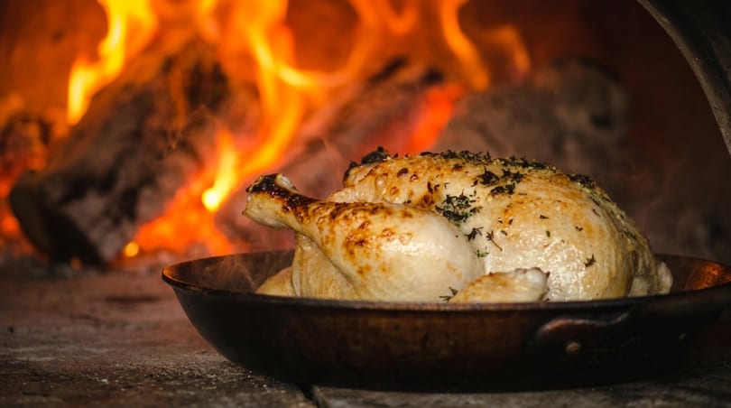 Whole roasted chicken in a pan, cooking in a wood-fired oven with visible flames.