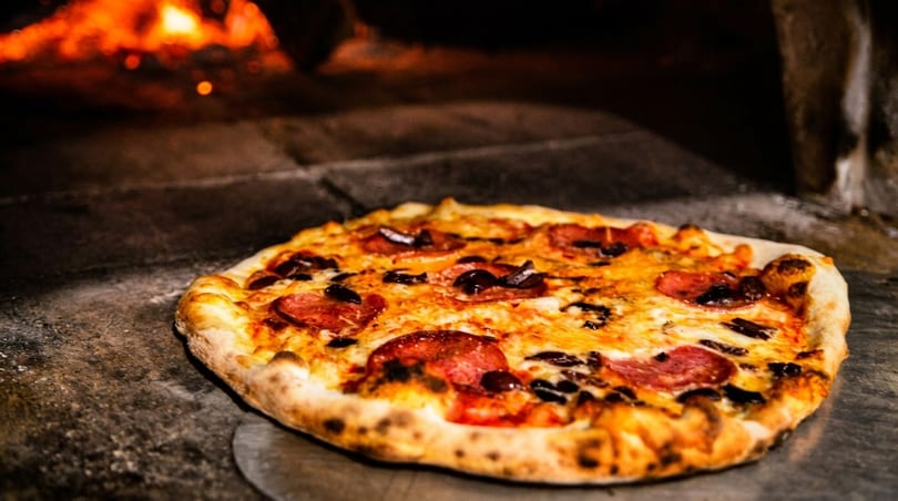 Freshly baked pepperoni and olive pizza on a peel, with a wood-fired oven in the background.
