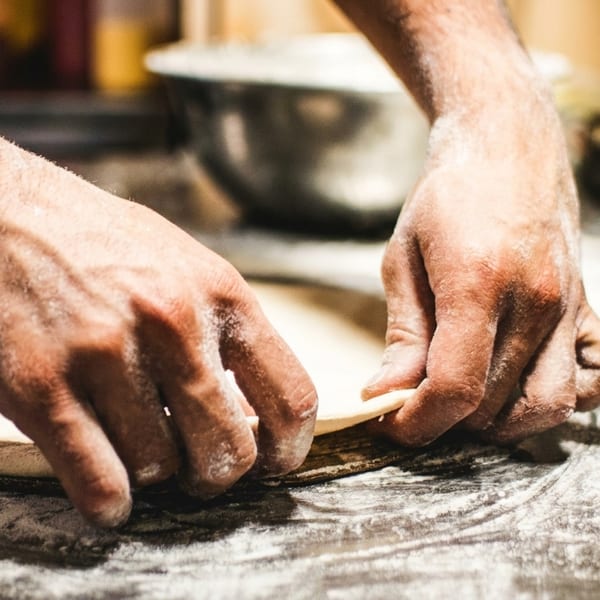 Hands covered in flour, shaping pizza dough on a dark, floured surface.