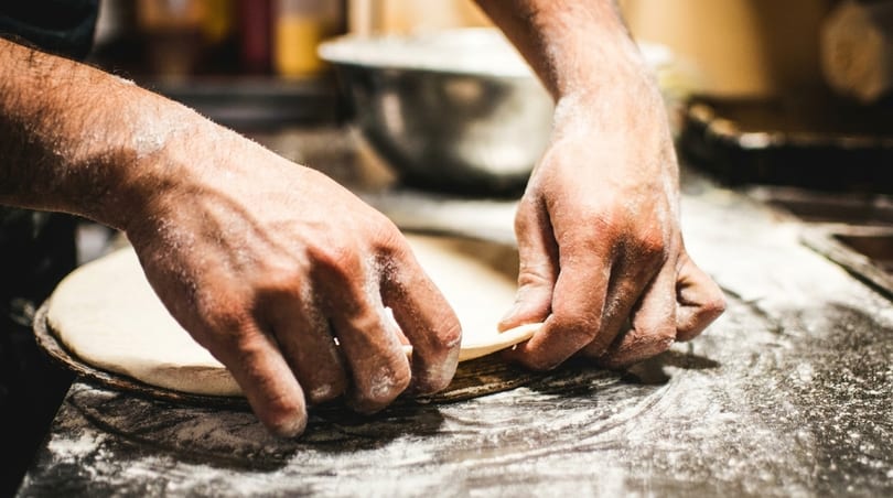 Hands covered in flour, shaping pizza dough on a dark, floured surface.